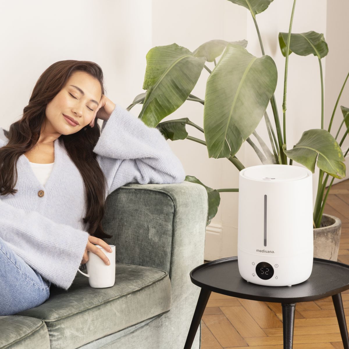 A humidifier stands on a small table next to a sofa and a houseplant while a woman relaxes holding a cup.